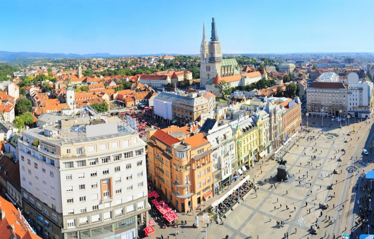 View from high above sunny Zagreb, where people are walking from across the main square to red umbrella covered markets